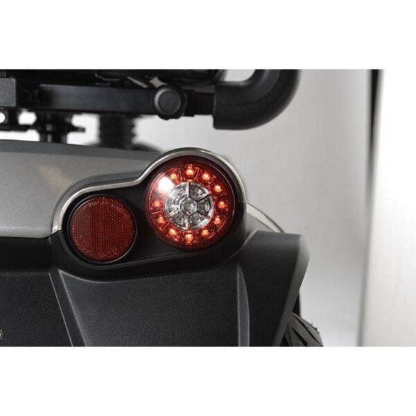 Close-up van het achterlicht van de Scootmobiel Kymco Maxer 4-wiel, met twee ronde LED-lampen. Het hoofdlicht schijnt rood met een metalen rand, gemonteerd op een strak grijs en zwart oppervlak.