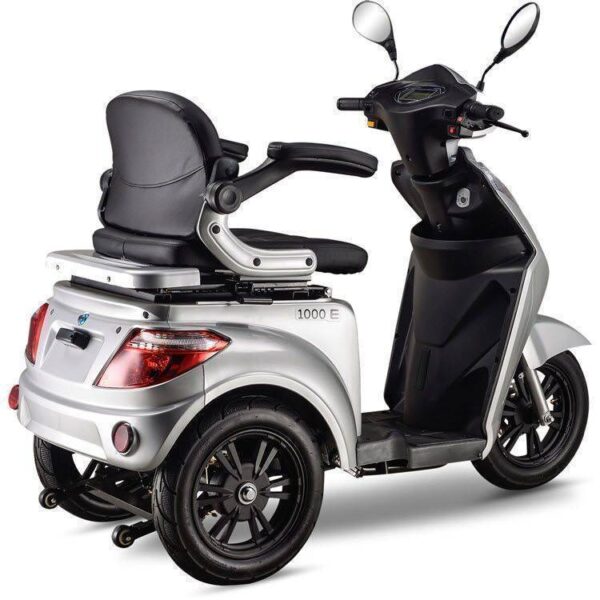 De IVA e1000 Scootmobiel is een zilveren driewielige elektrische scooter met een zwarte zitting en armleuningen, met achterstabilisatiewielen, verlichting en stuur met spiegels. Het modelnummer 1000 E is zichtbaar op de zijkant.